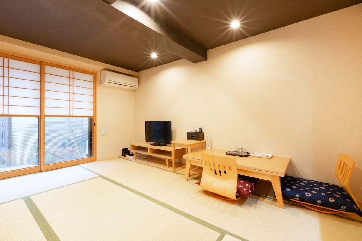 A spacious living area features tatami flooring and sliding shoji doors that invite natural light. A low wooden table is accompanied by two chairs and cushions, while a television and media setup are positioned nearby. Soft lighting enhances the calming atmosphere.
