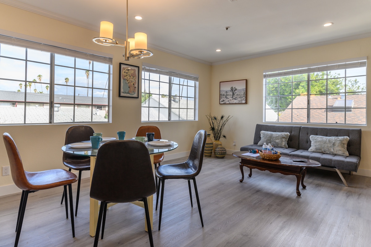 A well-lit living space features large windows allowing natural light to fill the room. A round dining table set for four stands beside an attractive coffee table. A comfortable sofa with cushions is positioned across from the dining area, enhancing the inviting atmosphere.