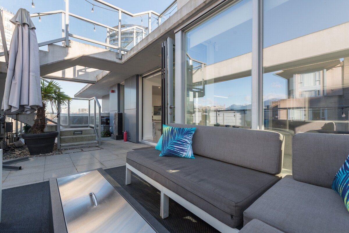 A modern outdoor space is shown, featuring a comfortable sectional sofa with decorative cushions. A fire table is positioned at the center, surrounded by stone pavers. A large umbrella provides shade, and potted plants add a refreshing touch. Views of the surrounding cityscape can be glimpsed through the glass doors.