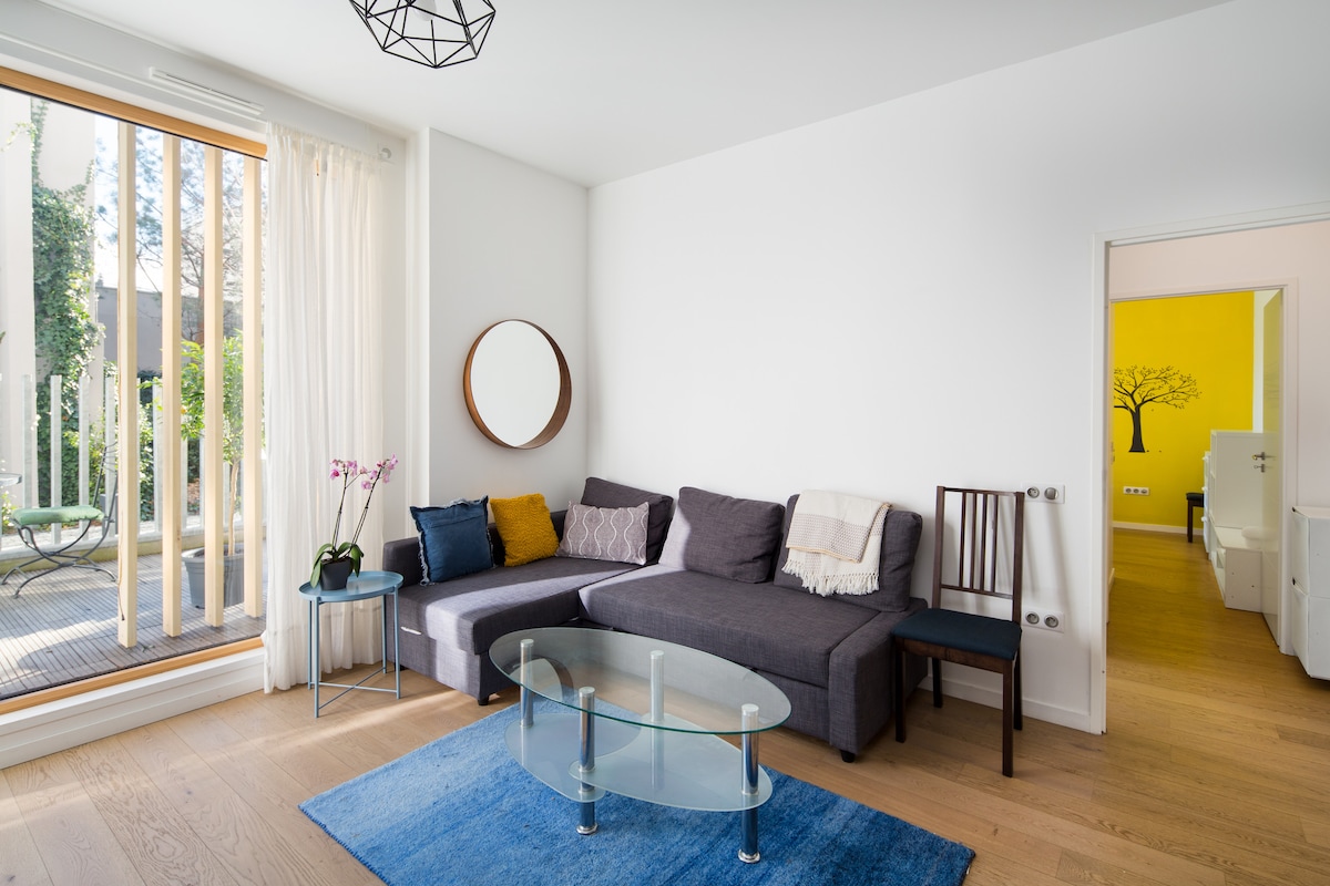 A spacious living area features a gray sectional sofa with colorful cushions, a round glass coffee table, and a large mirror. Natural light enters through tall glass doors leading to a private terrace. A vibrant yellow wall is visible in the adjoining room, providing a cheerful contrast.