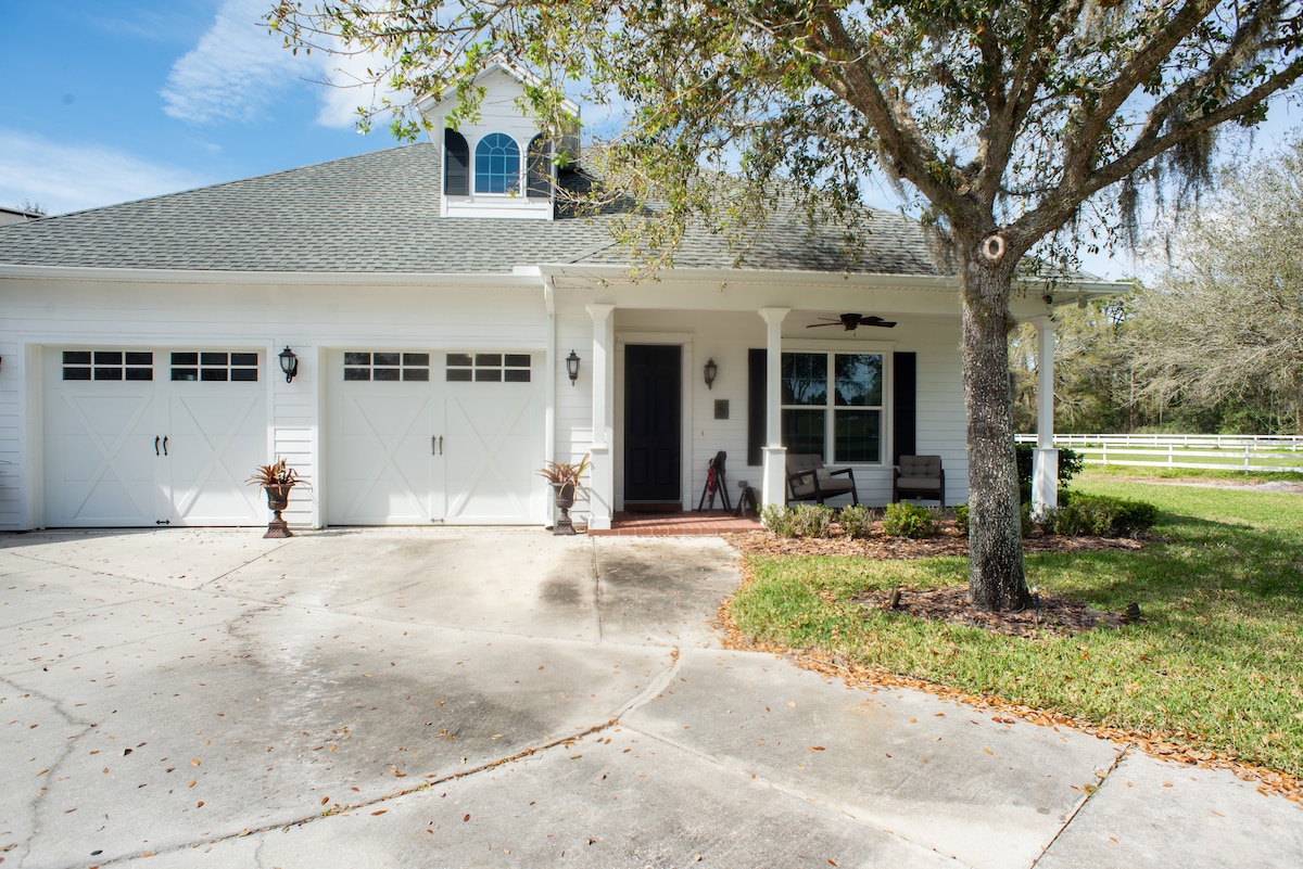 The exterior of the cozy apartment cottage is shown, featuring a welcoming front porch with two comfortable chairs. A well-maintained driveway leads to the two-car garage, surrounded by lush green grass and trees, offering a serene atmosphere near the entryway.