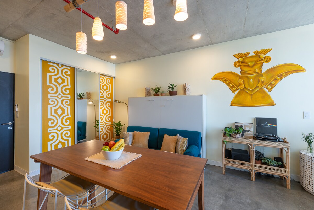 A modern living space features a blue sofa paired with decorative cushions, positioned near a wooden dining table with a fruit centerpiece. Unique wall art inspired by indigenous culture adds vibrancy, while warm lighting from hanging fixtures creates a cozy ambiance. A mirrored closet reflects the design.