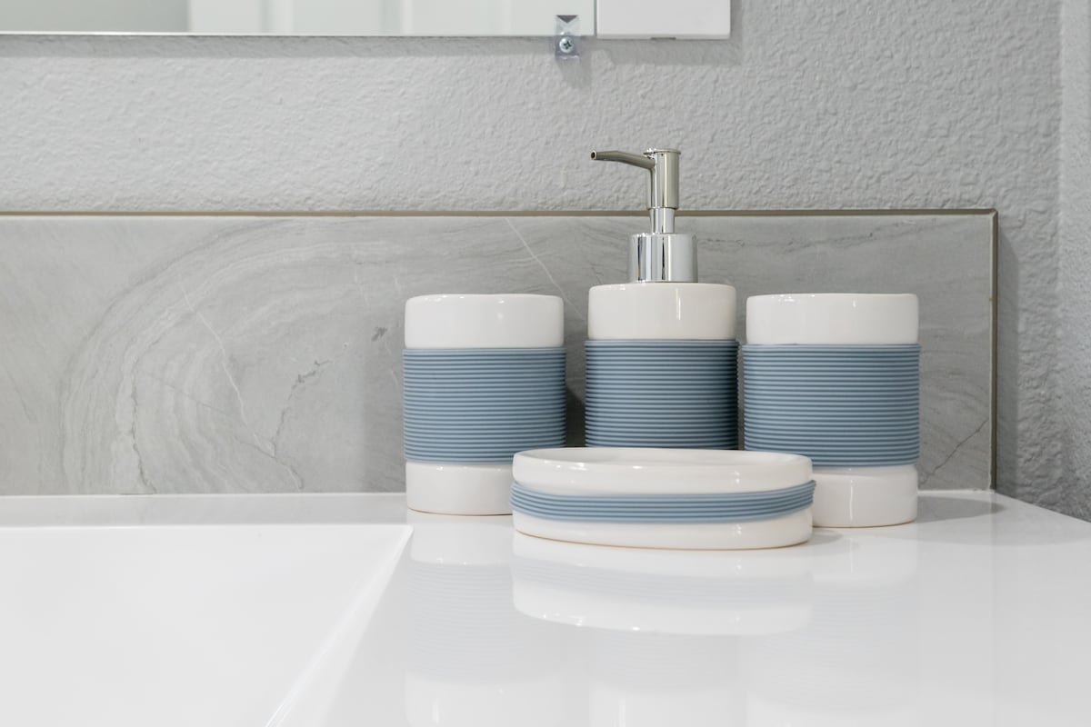 A modern bathroom countertop is displayed, featuring a polished white surface. Three coordinating container pieces with a calming blue band are arranged, including a soap dispenser and two matching containers, alongside a dish for small items. A mirror is positioned above the countertop.