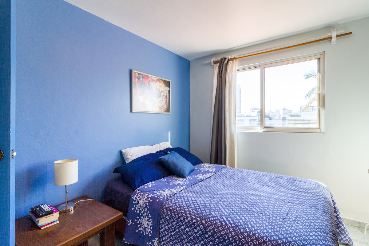 A tranquil bedroom features soft blue walls and natural light coming through a large window. A neatly made bed with a blue patterned duvet is positioned beside a small nightstand, which holds a lamp and several books. A piece of art is displayed above the bed.