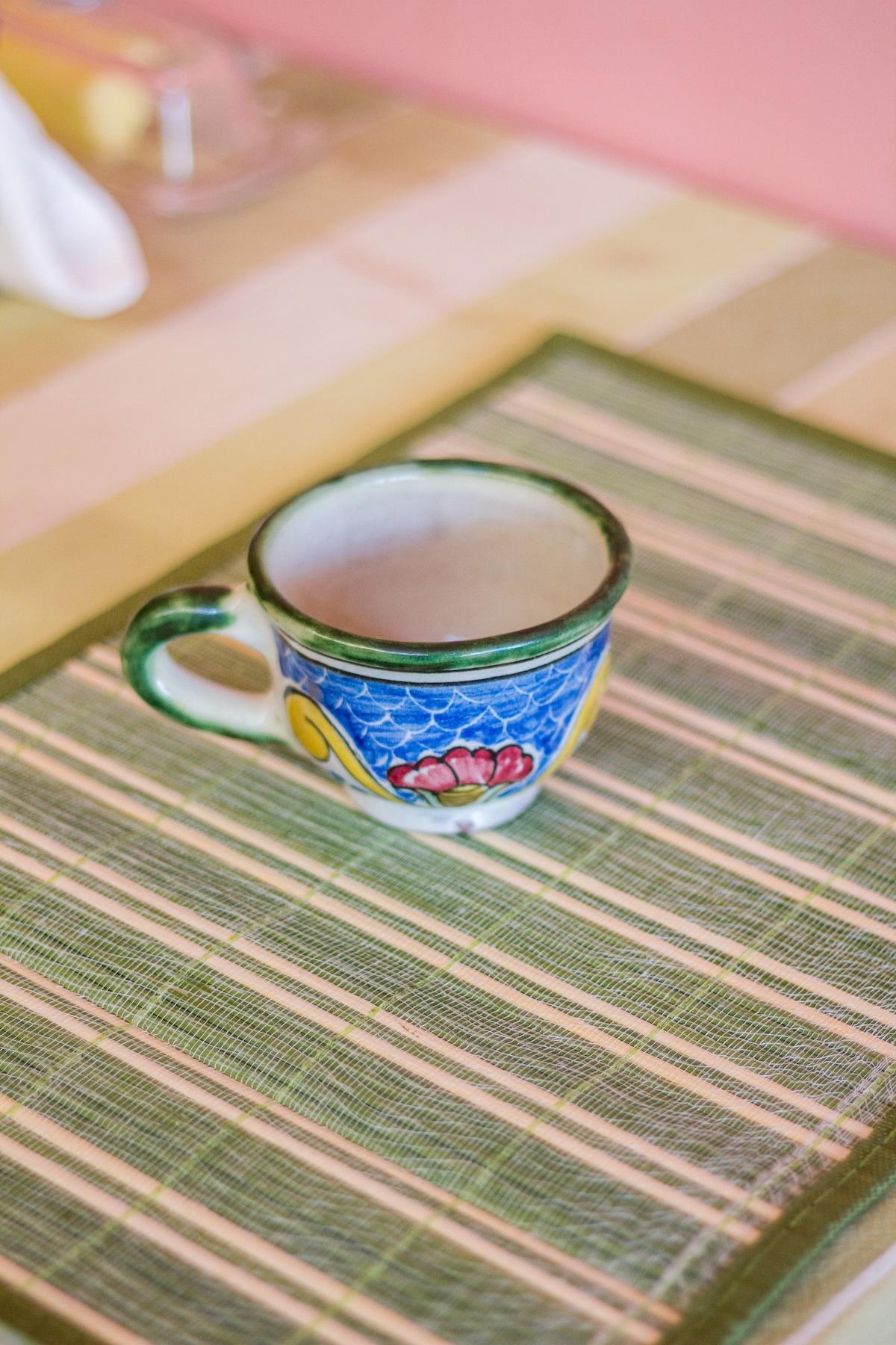 A colorful ceramic mug is placed on a striped green placemat, featuring a floral design and a sturdy handle. The mug's vibrant colors complement the decor and add a touch of charm to the setting.
