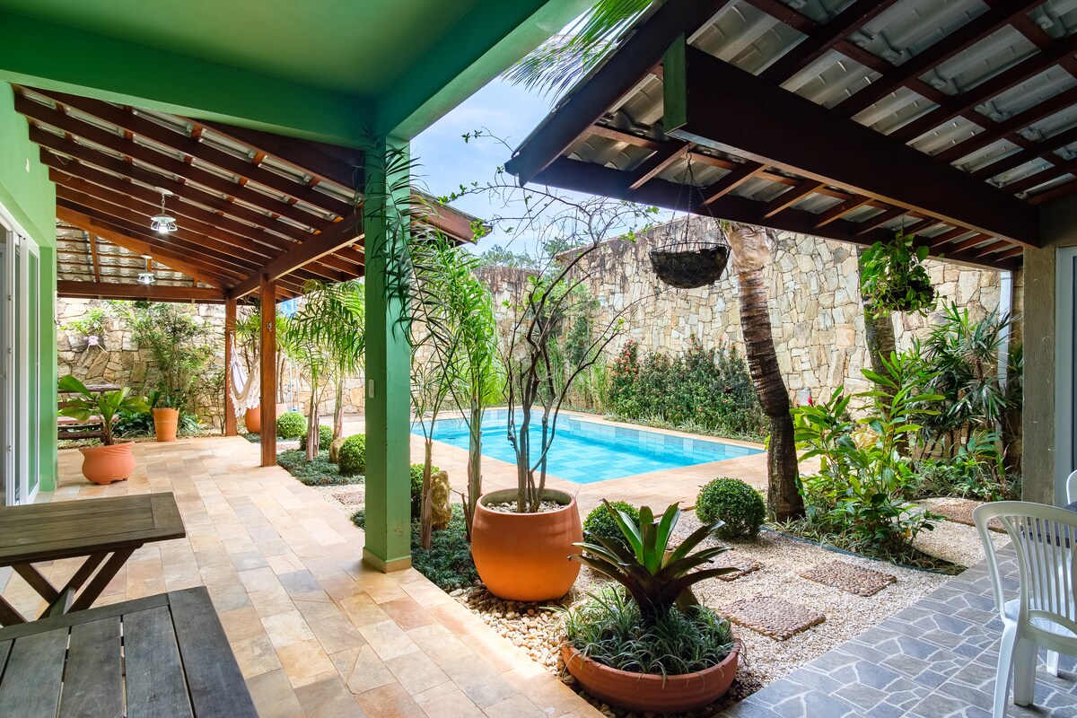 An outdoor living area features a covered patio with wooden beams and a large dining table. Lush greenery surrounds a pool, complemented by decorative pots and stone pathways.