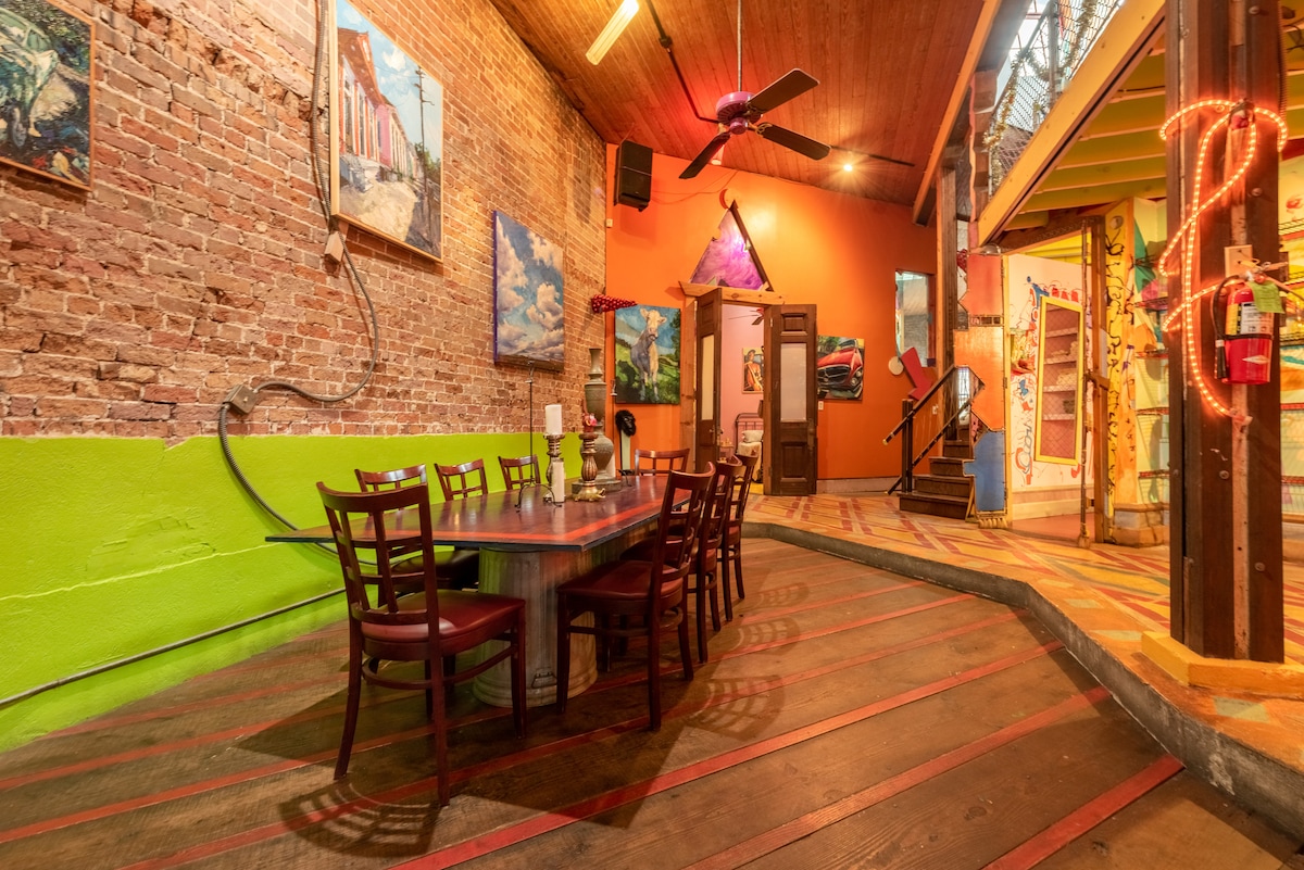 A vibrant dining area is highlighted by a long wooden table surrounded by several chairs. The exposed brick walls are adorned with colorful artwork, and the space features lively orange and green accents. Unique flooring leads towards an open doorway, inviting exploration.