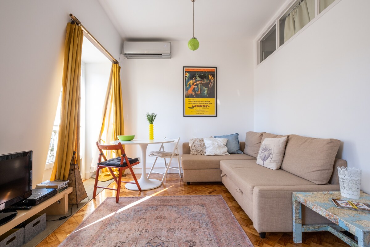 A bright living area features a comfortable sofa and a small dining table with two chairs. Large windows adorned with flowing yellow curtains let in ample light, creating a warm ambiance. A vintage poster is displayed on the wall, enhancing the space's character.