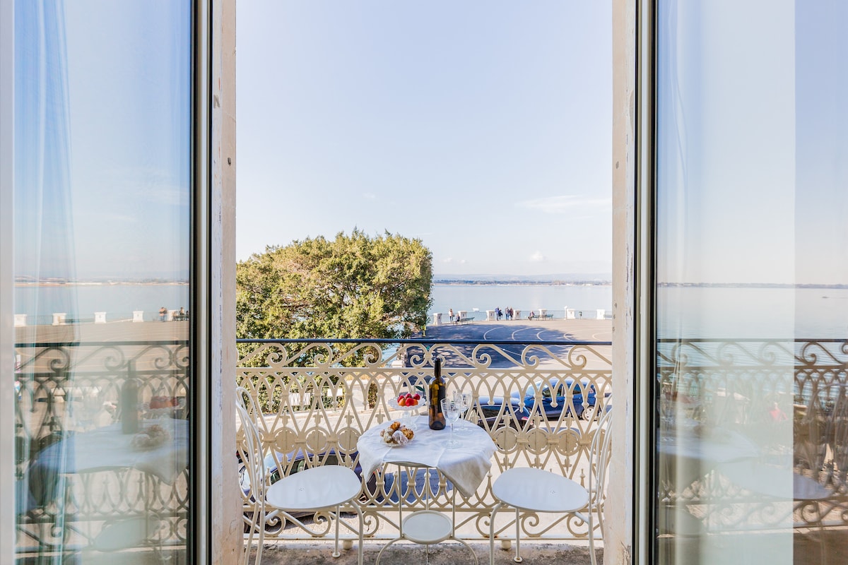 A balcony overlooks the water, featuring elegant wrought-iron railings. A small table is set with two chairs, inviting relaxation. The panoramic view includes a glimpse of the waterfront and trees, creating a serene atmosphere.