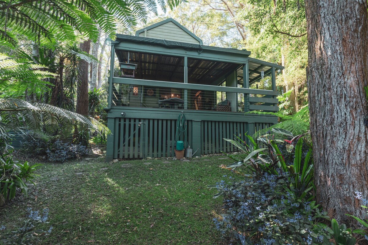 The cottage is framed by lush greenery, showcasing a timber structure with a covered porch and surrounding foliage. The vibrant garden features various plants, creating a serene atmosphere. A gentle slope leads to the entrance, enhancing the cabin's cozy, private setting.