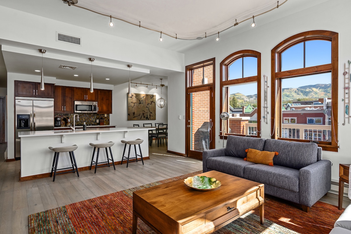 A bright and airy living area features a gray sofa facing a wooden coffee table adorned with a colorful rug. Behind, a kitchen area with modern appliances and an island is complemented by windows offering scenic views of the surrounding mountains.
