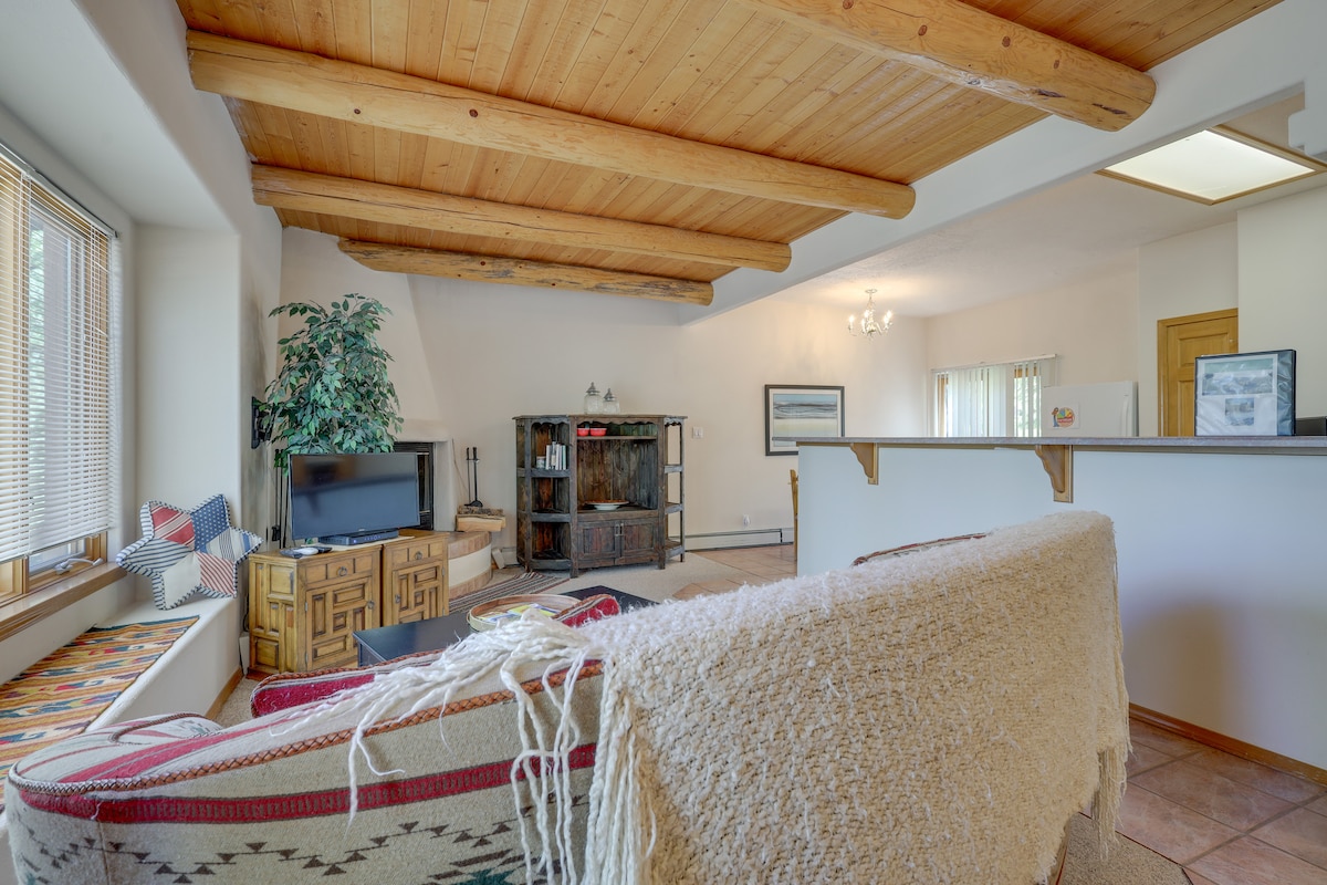A spacious living area features wooden beams across the ceiling and a cozy seating area with a patterned blanket draped over a sofa. A television is positioned on a wooden stand, while a decorative shelf adds interest. Natural light enters through the wide window with a built-in seat.