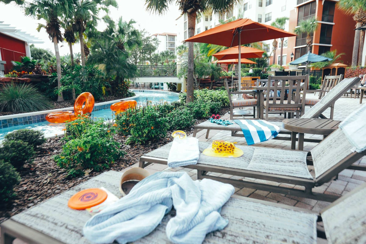 Loungers are arranged beside a landscaped pool area, surrounded by palm trees and bright orange umbrellas. Some pool toys and towels are scattered on the loungers, suggesting areas for relaxation and leisure. Lush greenery and a winding pool feature are visible in the background.