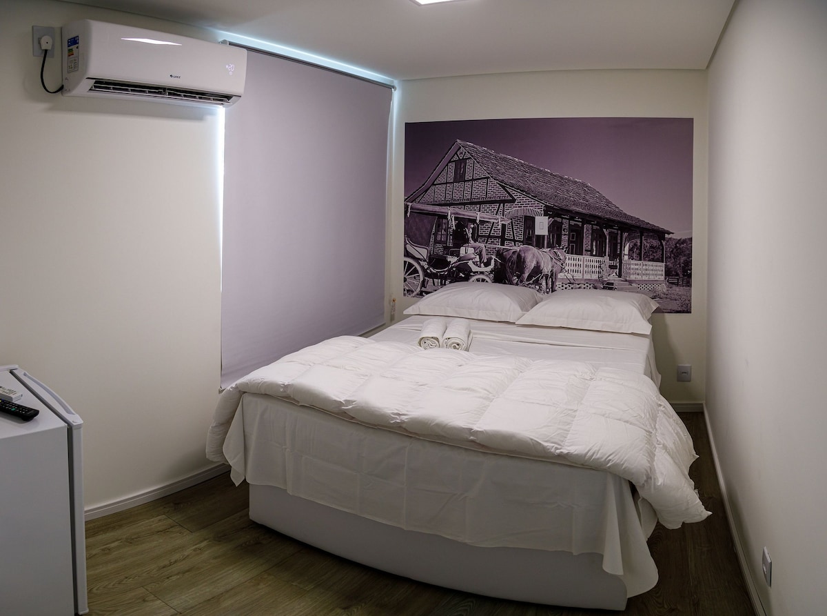 A cozy sleeping area features a spacious bed dressed in white linens, accompanied by neatly arranged towels. A large black-and-white photo of a rustic house adorns one wall, while a window with a roller shade provides natural light. An air conditioning unit is visible above.