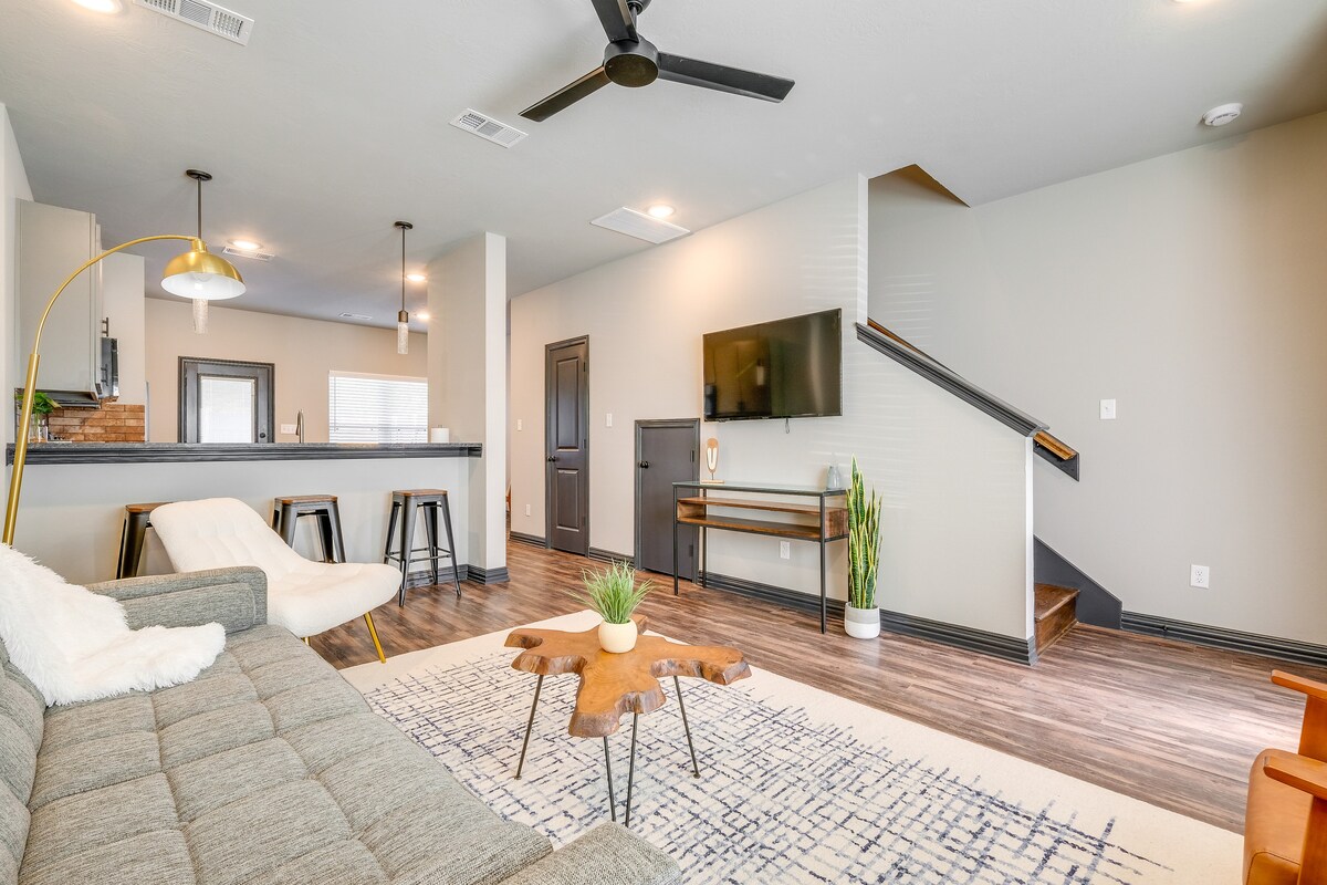 A contemporary living area features a comfortable sectional sofa and a stylish coffee table at its center. A mounted Smart TV is positioned on the wall, and a set of bar stools complements the nearby kitchen. Natural light is provided by large windows, and a ceiling fan is present for airflow.
