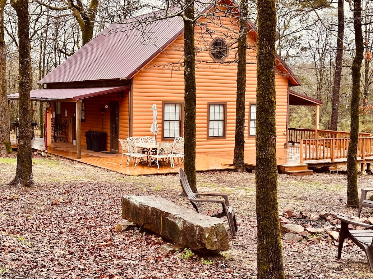 The rustic log cabin features a warm wood exterior with a dark metal roof. A spacious deck is visible, furnished with a dining table and chairs, surrounded by tall trees. A fire pit and seating area are located in the foreground, creating a relaxed outdoor space.
