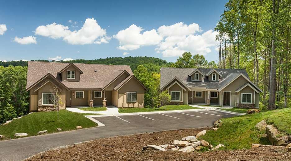 Two charming lodges are showcased under a blue sky, surrounded by lush greenery. The buildings feature light-colored exteriors with stone accents. A paved driveway leads to both structures, arranged side-by-side against a backdrop of rolling hills and trees.