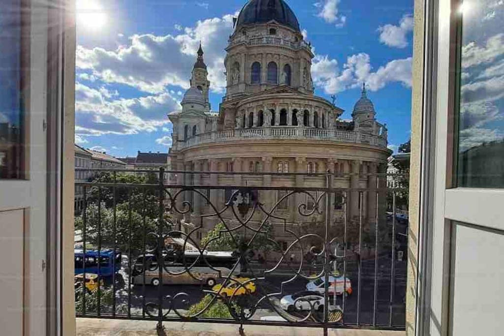 A balcony view showcases the grand architecture of St. Stephen's Basilica. The ornate dome and intricate details are framed by the open balcony doors, while a vibrant sky partially covered by clouds adds depth to the scene. Streets below reveal vehicles and greenery.