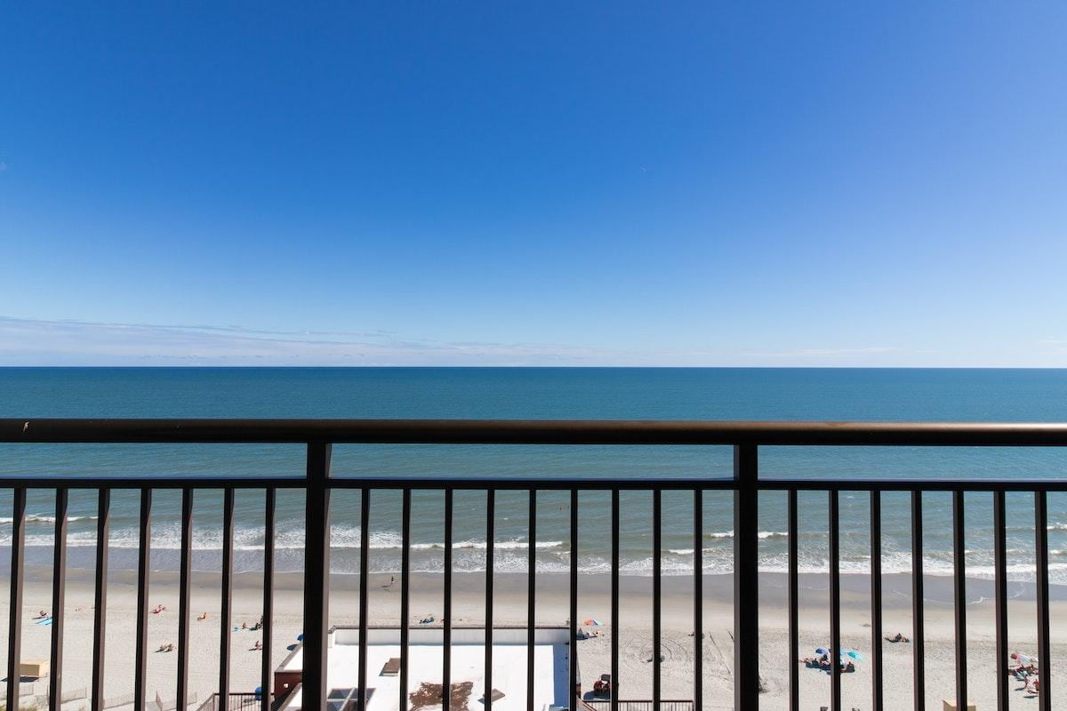 A private oceanfront balcony offers unobstructed views of the shoreline and the Atlantic Ocean. The clear blue sky reflects over the water, while beachgoers can be seen along the sandy shore. The warm tones of the beach contrast with the vibrant colors of the ocean.
