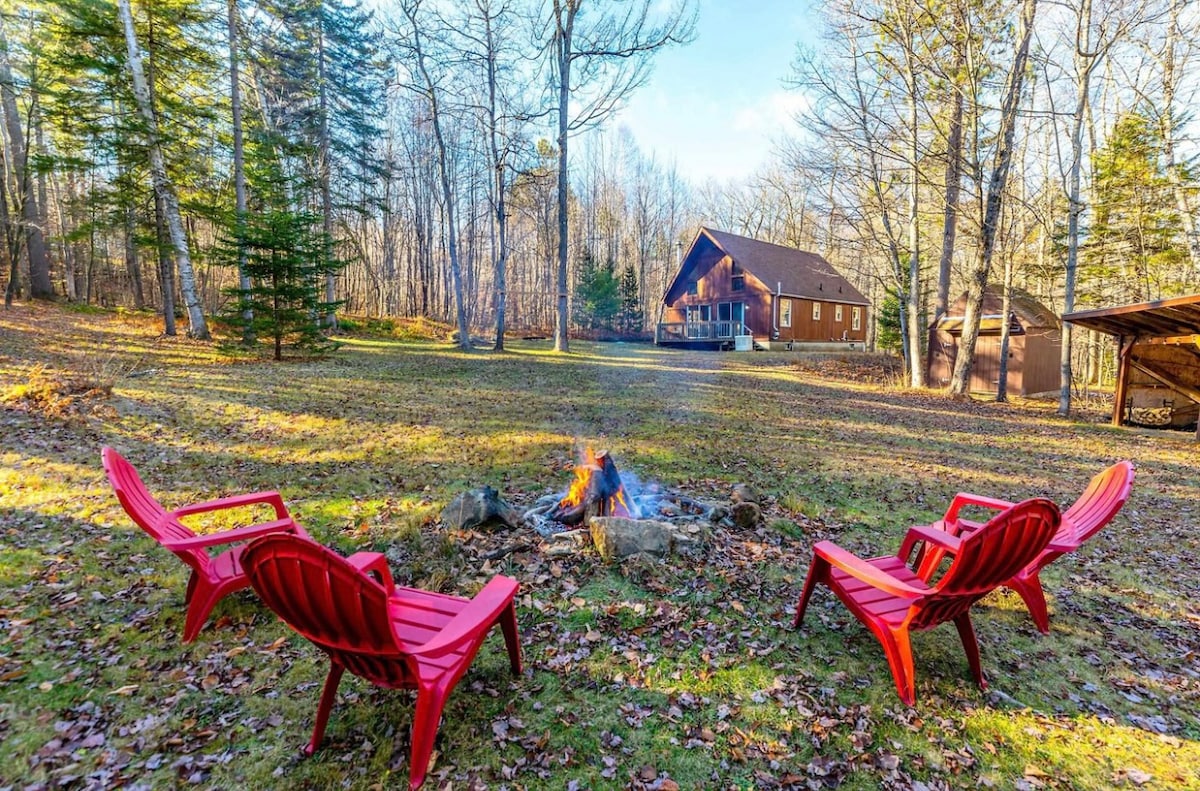 Two red Adirondack chairs are positioned around a fire pit in a peaceful outdoor setting. The background features a spacious cabin surrounded by tall trees and a grassy area, creating an inviting atmosphere for relaxation and gatherings.