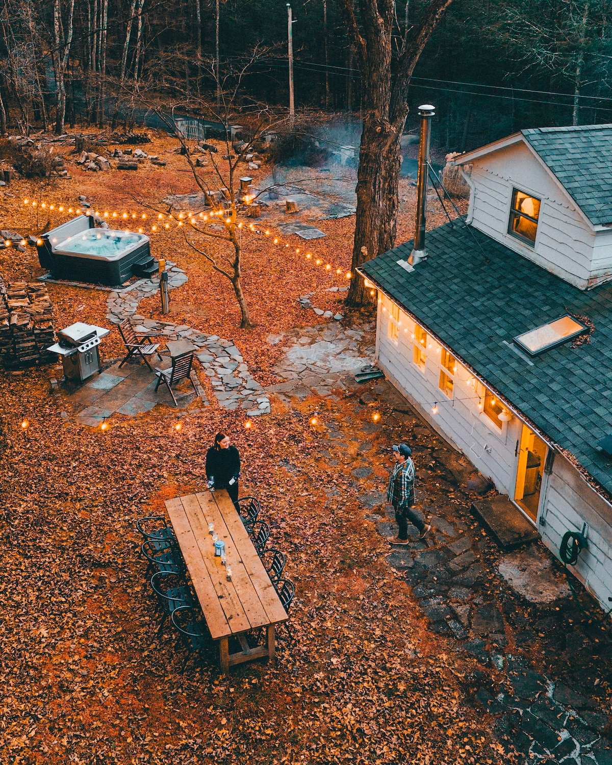 An aerial view of an outdoor space reveals a rustic cabin surrounded by a carpet of fall leaves. A large wooden dining table is set with chairs, while string lights create a warm atmosphere. A hot tub and fire pit invite guests to enjoy the natural setting.