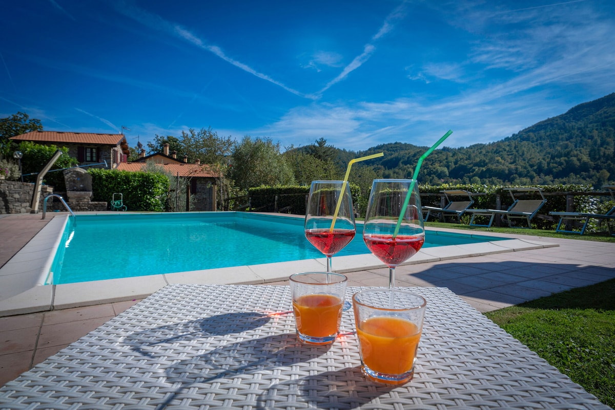 The outdoor pool area is presented, featuring a heated swimming pool surrounded by lounge chairs. Three drinks are placed on a white wicker table, with two cocktails in glasses and one orange beverage. The backdrop includes scenic greenery and mountains under a clear blue sky.