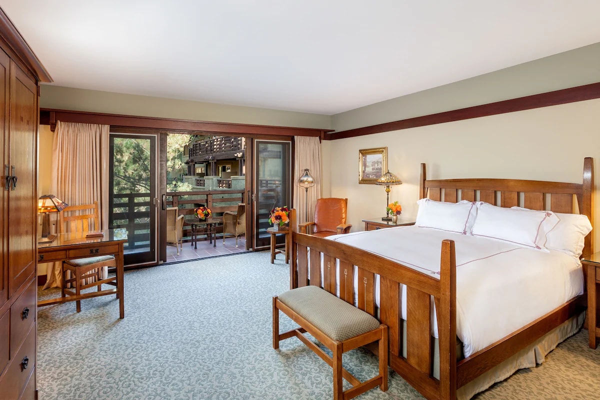 A spacious guest room features a wooden bed with crisp linens and decorative pillows. Natural light enters through large sliding doors, leading to a private balcony. A small seating area includes a wooden table and chair, with a dresser and additional seating visible.