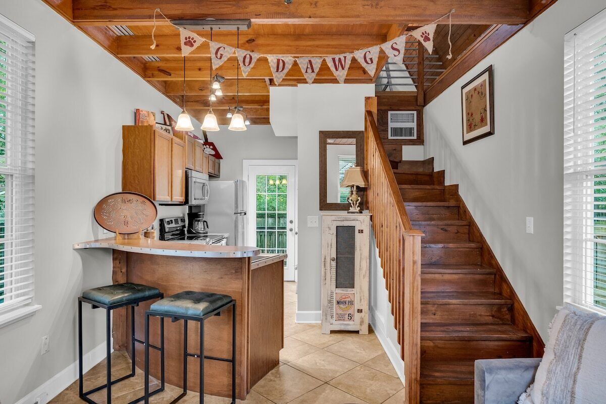 A cozy kitchen area features warm wooden cabinetry and a decorative banner reading 'GO DAWGS' overhead. Two bar stools are placed at a counter, with steps leading to the upper level in the background. Natural light enters through large windows, creating an inviting ambiance.