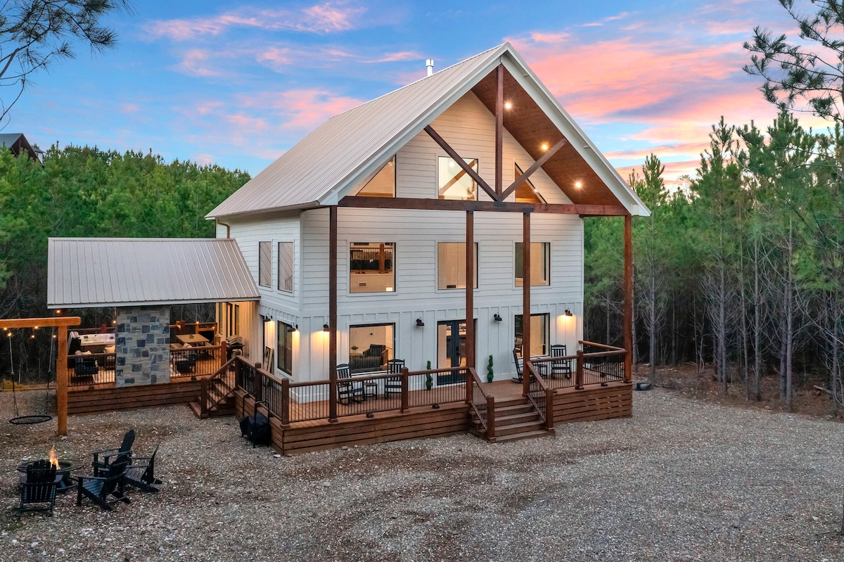 A modern rustic cabin is set against a backdrop of trees and a colorful sunset. The exterior features large windows with wooden accents, and a spacious deck furnished with seating. An outdoor gathering area with chairs and a nearby grill is visible on the side.