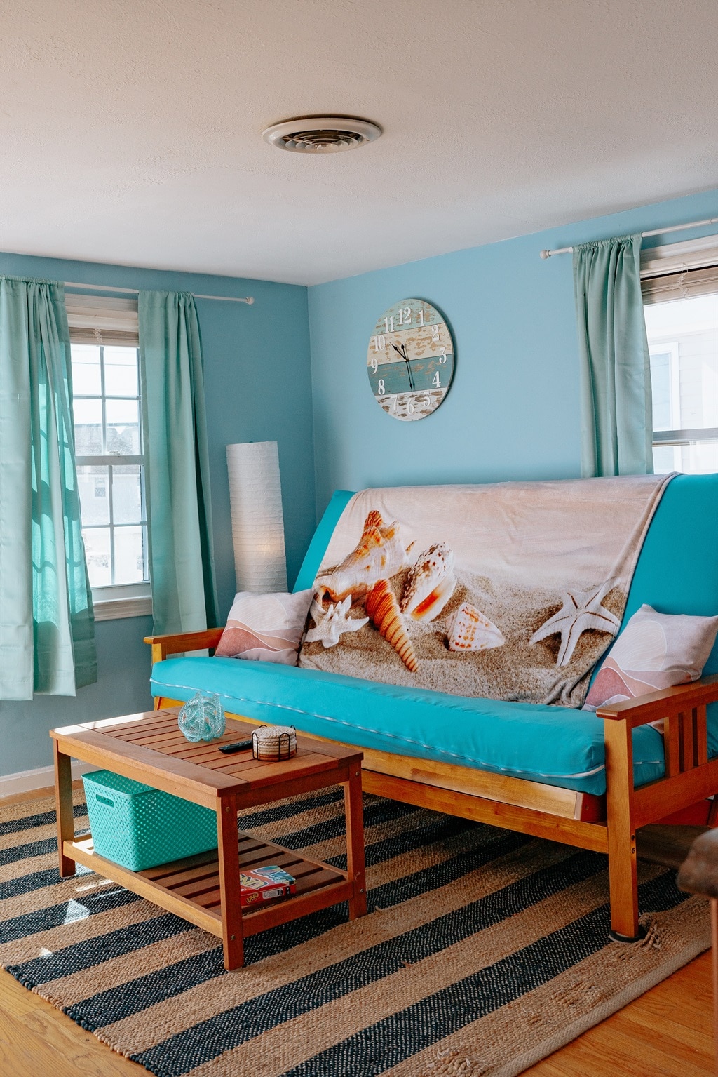 A cozy living area features a convertible futon adorned with a beach-themed blanket. Light blue walls are complemented by bright curtains and natural light streaming through the windows. A small wooden table sits on a striped rug, adding warmth to the space.