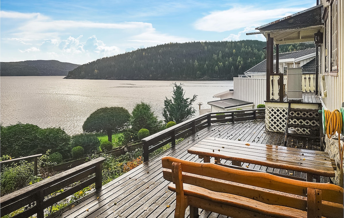 A wooden terrace features several bench seats overlooking a vast body of water. Lush green hills are seen in the distance, and a few trees line the terrace area, creating a natural backdrop against the sky.