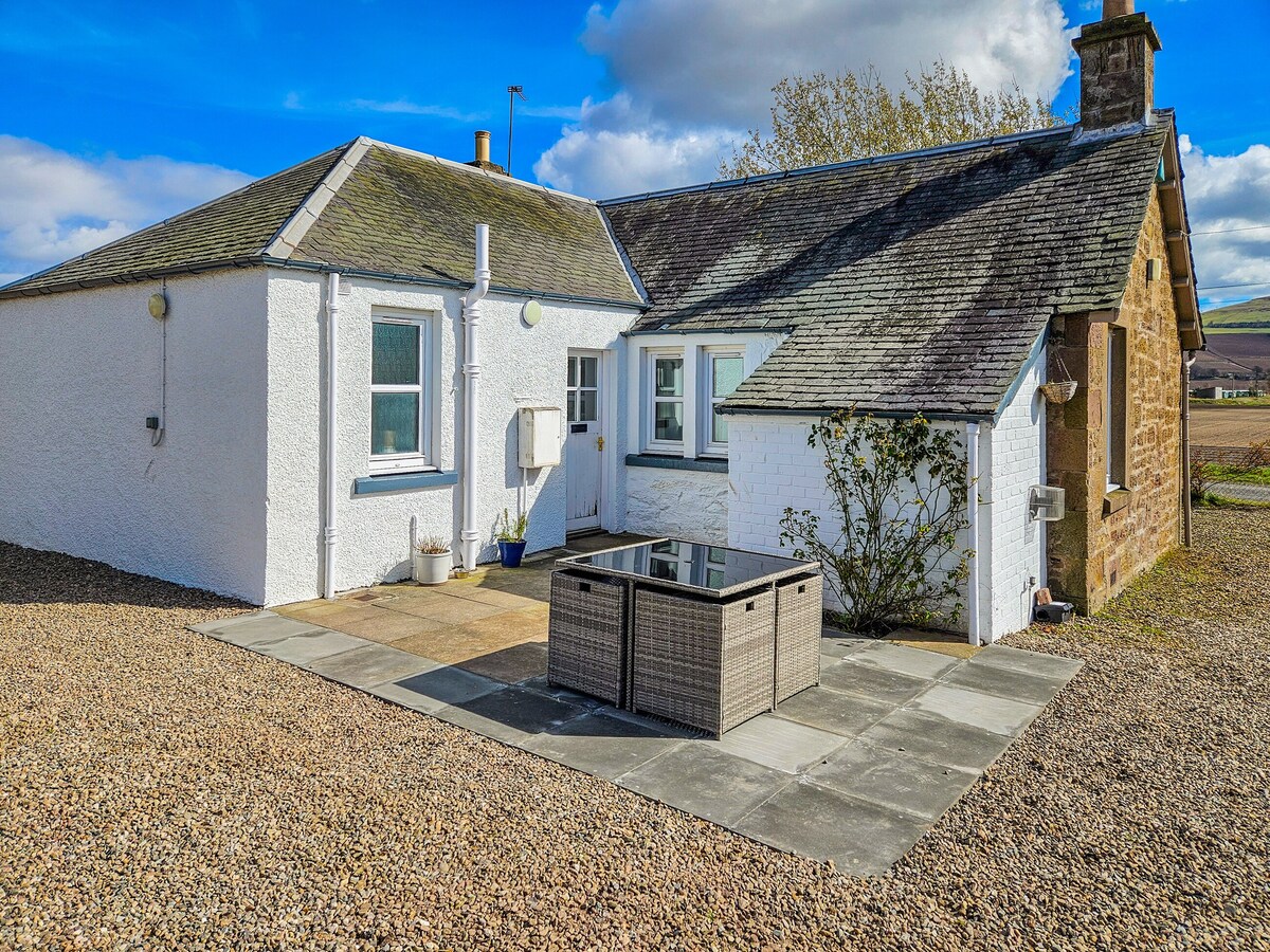 A charming cottage exterior is set against a clear blue sky, showcasing a spacious stone patio. A modern outdoor table with glass top sits at the center, surrounded by light gravel and landscaped elements. Nearby, a mix of traditional and modern architecture is visible.