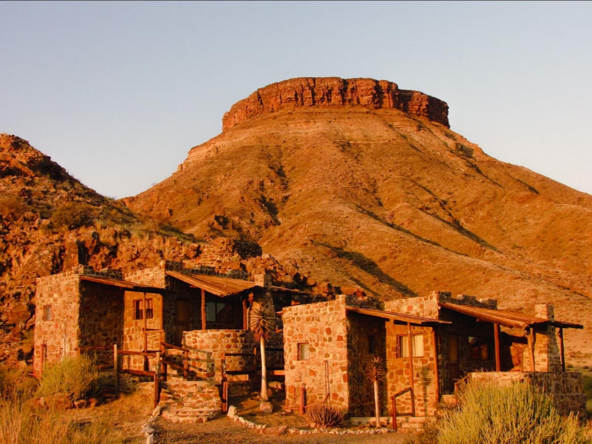 The rustic stone structure is nestled against a backdrop of a large, flat-topped mountain. The warm earth tones of the building complement the surrounding rocky landscape, while patches of green vegetation can be seen in the foreground.