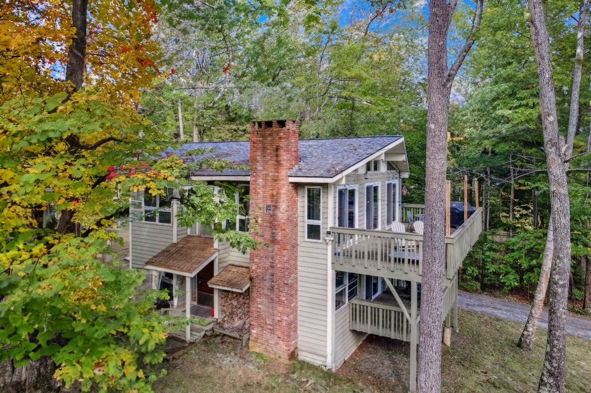 The vacation home features a multi-level structure nestled among trees, showcasing a rustic brick chimney and expansive windows. A spacious deck extends from the upper level, offering a view of the surrounding nature. The exterior is complemented by vibrant autumn foliage.