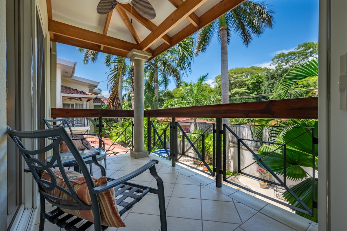 A terrace features two comfortable chairs with decorative cushions, positioned to take in views of lush tropical greenery. Ceiling fans provide a cooling breeze under the wooden beam roof, creating a serene space for relaxation in a tranquil outdoor setting.