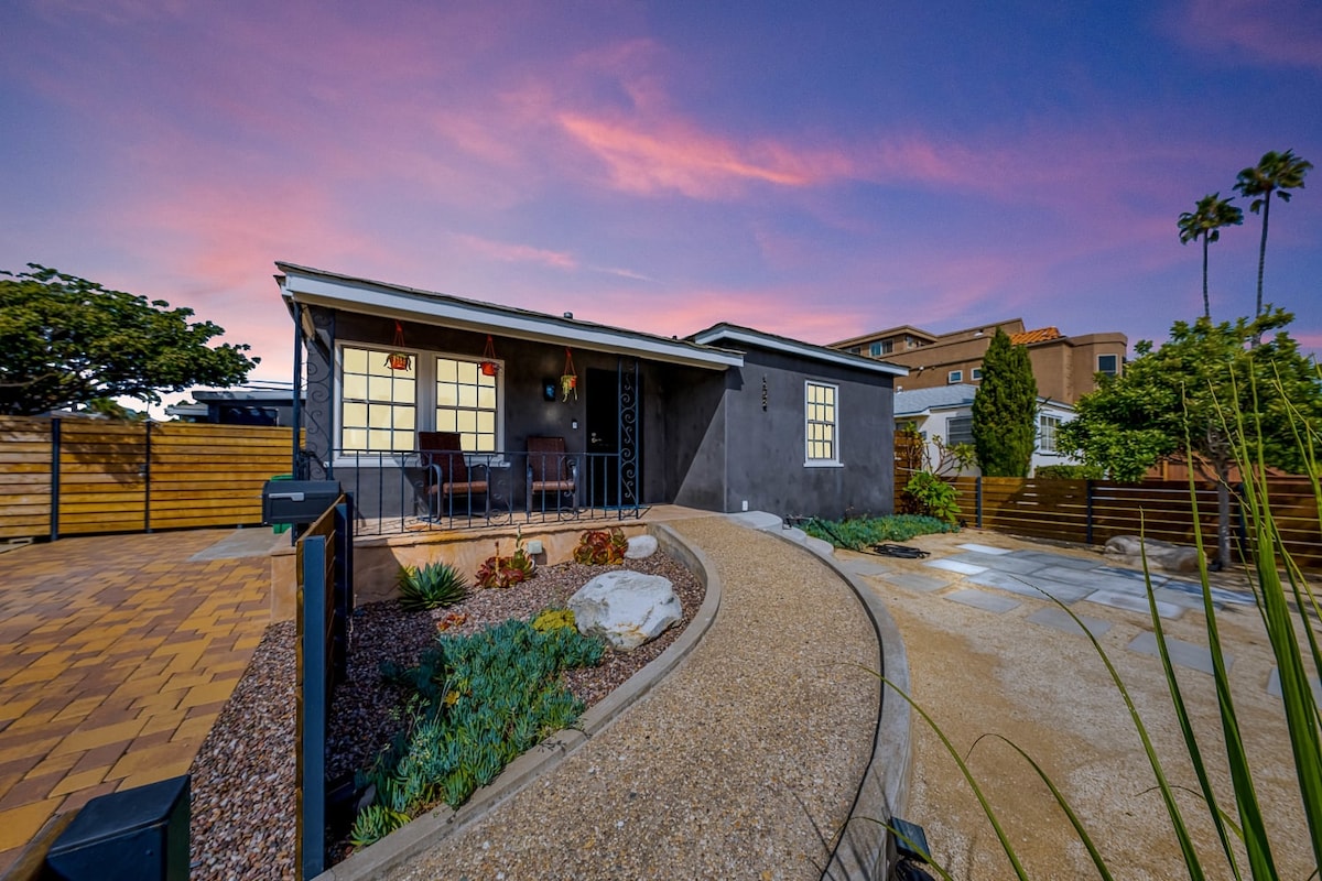 The exterior of the property is highlighted by a modern facade, showcasing a well-maintained garden with a winding pathway. Two chairs are positioned on the porch, inviting relaxation while enjoying the colorful sunset sky and surrounding tropical landscaping.
