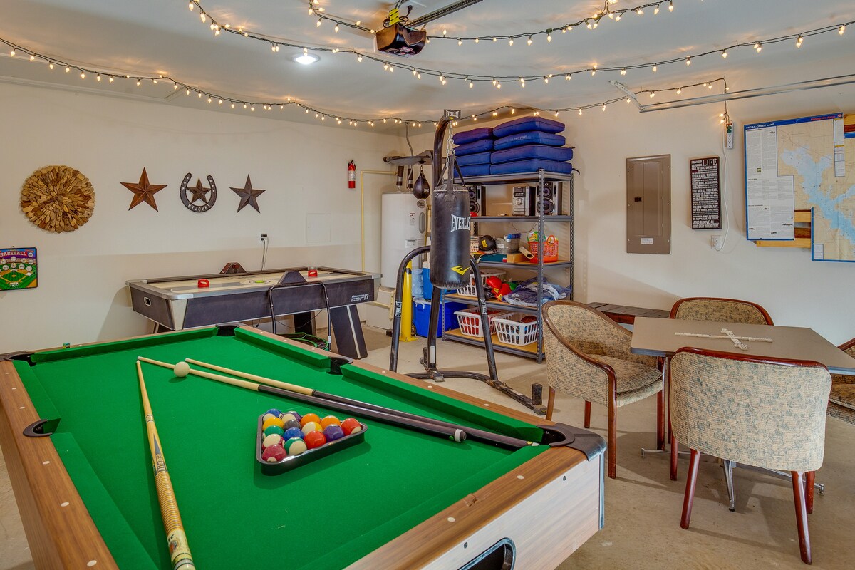 A spacious game room is equipped with a pool table featuring a triangle of colored balls. A table and chairs are available for seating, while an air hockey table is positioned nearby. Strings of lights decorate the ceiling, adding a relaxed ambiance.