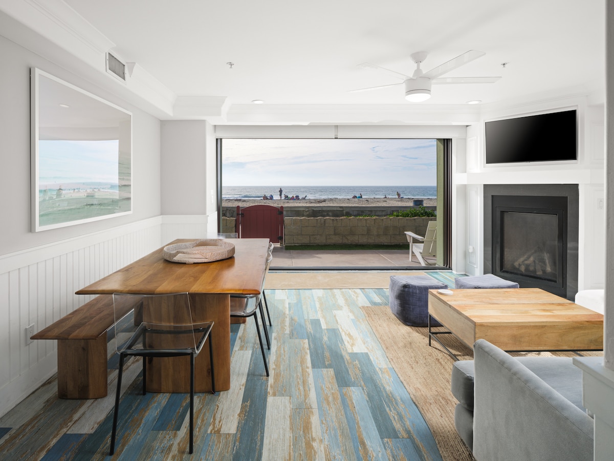 A spacious living area is highlighted by a large dining table and comfortable seating. Expansive glass doors open to reveal ocean views, allowing natural light to illuminate the room. The area is complemented by a cozy fireplace and thoughtfully chosen decor elements.