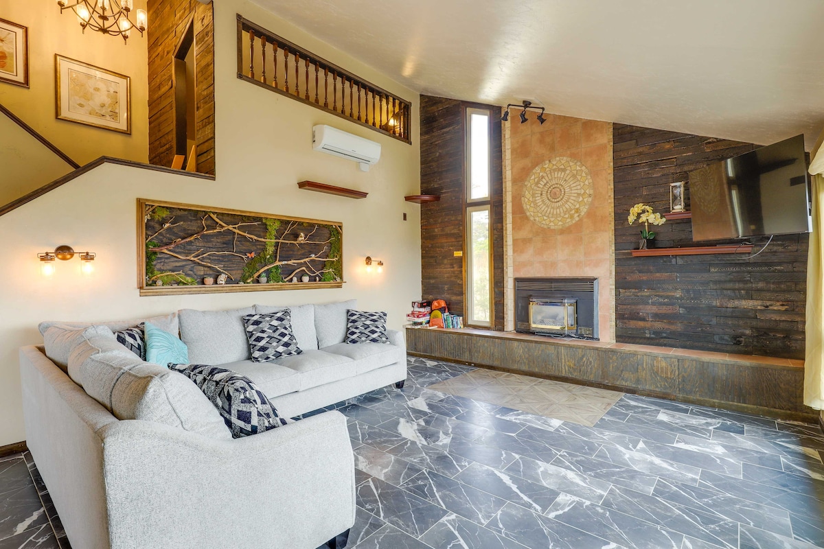 A spacious living area features a gray sectional sofa positioned near a modern gas fireplace. Large windows allow natural light to fill the room, highlighting wooden accents on the walls. A staircase leads to the upper level, enhancing the home's verticality.