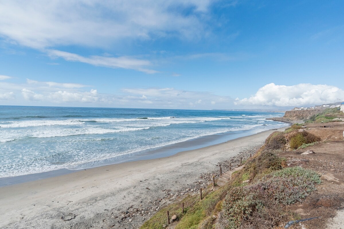 A scenic view of the coastline showcases gentle waves lapping at a sandy beach. The water reflects a clear blue sky, while patches of greenery line the shore, contributing to the tranquil atmosphere. Clouds create a soft contrast against the vibrant horizon.