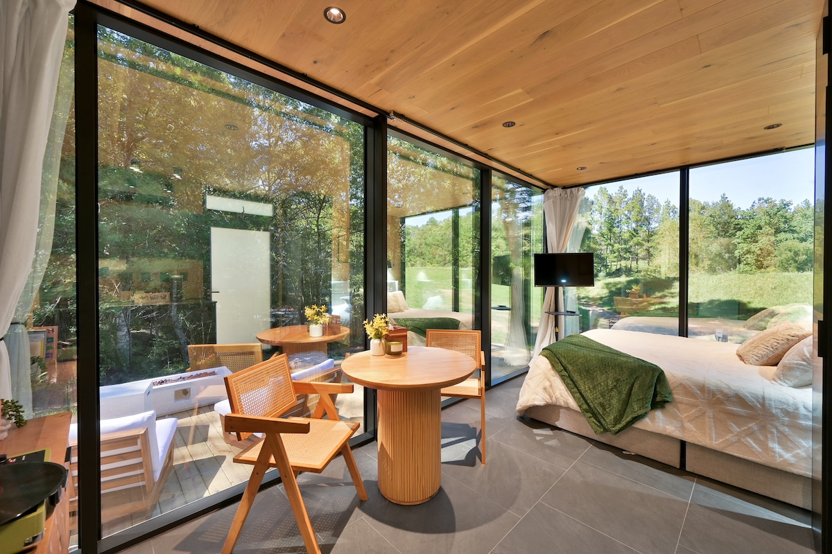 A spacious interior features floor-to-ceiling glass walls allowing natural light to fill the room. A comfortable bed is positioned near the window, while a wooden dining table and two chairs create a cozy seating area. A television is mounted on the wall for entertainment.
