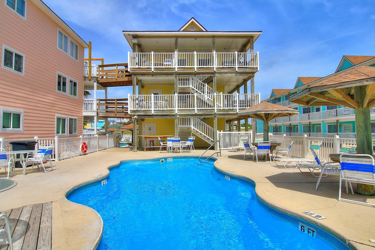 The image captures a central outdoor pool area, surrounded by a variety of lounge chairs and shaded by umbrellas. Adjacent buildings feature multiple balconies and light-colored exteriors, creating a relaxed environment. A spacious deck area allows for easy access to the pool and seating options.