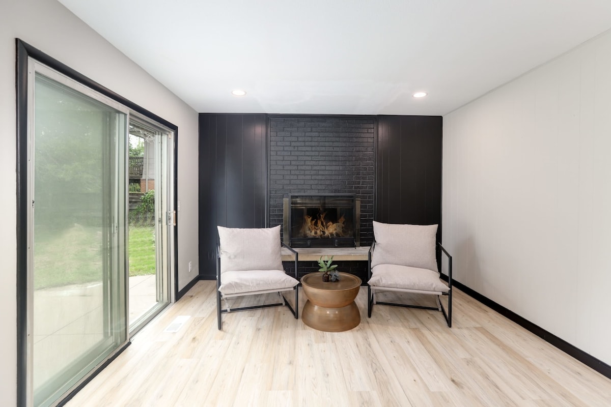 A cozy seating area features two light-colored chairs flanking a round table. A fireplace with a dark brick surround is centered on the wall. A sliding glass door leads to a green outdoor space, allowing for natural light to fill the room.