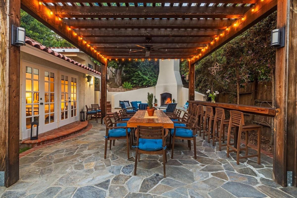 A spacious outdoor patio area is covered by a wooden trellis adorned with string lights. A large table with comfortable blue seating surrounds the space, creating an inviting atmosphere. A fireplace is visible in the background, adding warmth to the setting.
