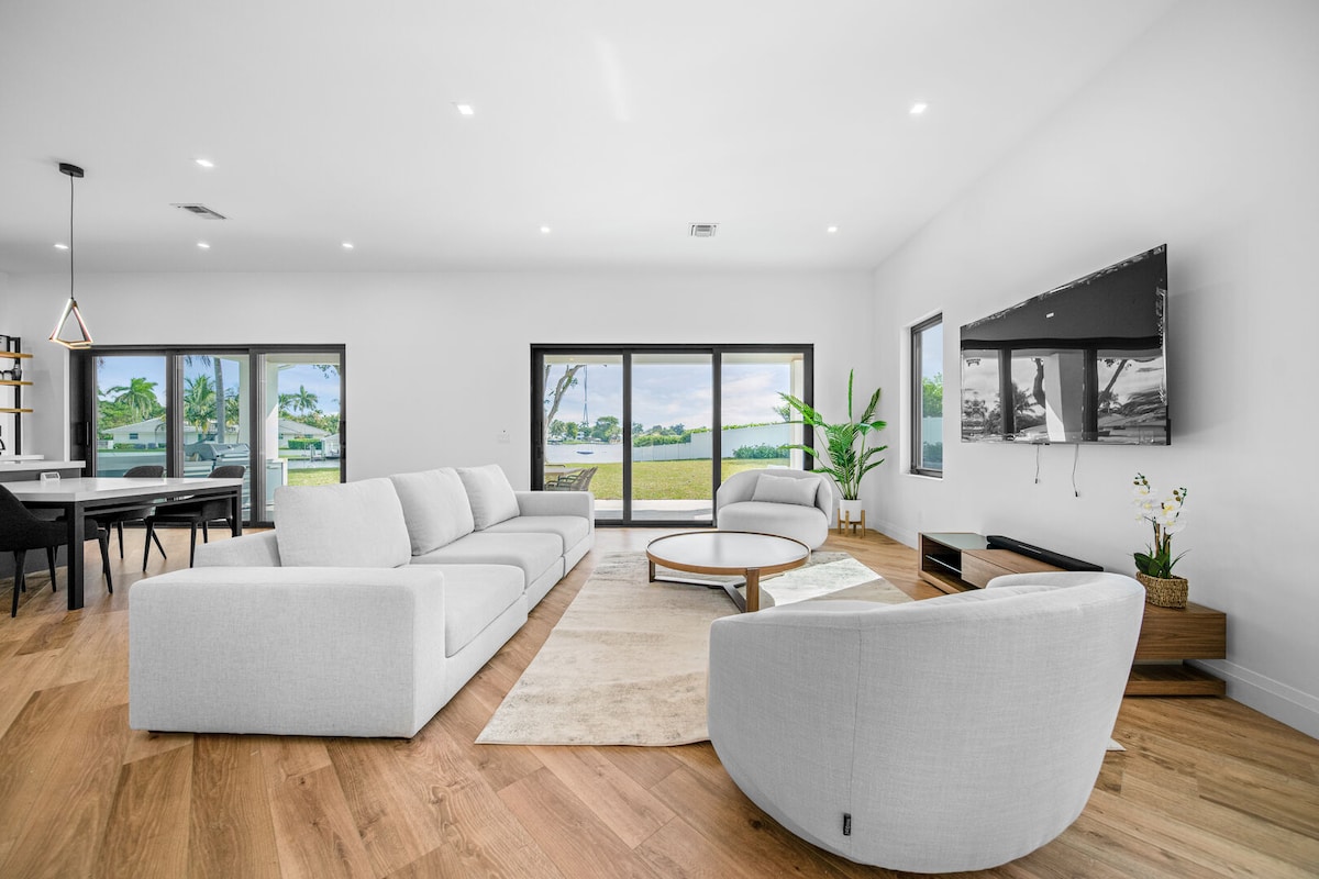 A spacious living area is furnished with a plush sectional sofa and a round coffee table, providing a comfortable space to relax. Large glass doors open to scenic views of the outdoors, while natural light fills the room. A sleek TV and modern decor complete the ambiance.