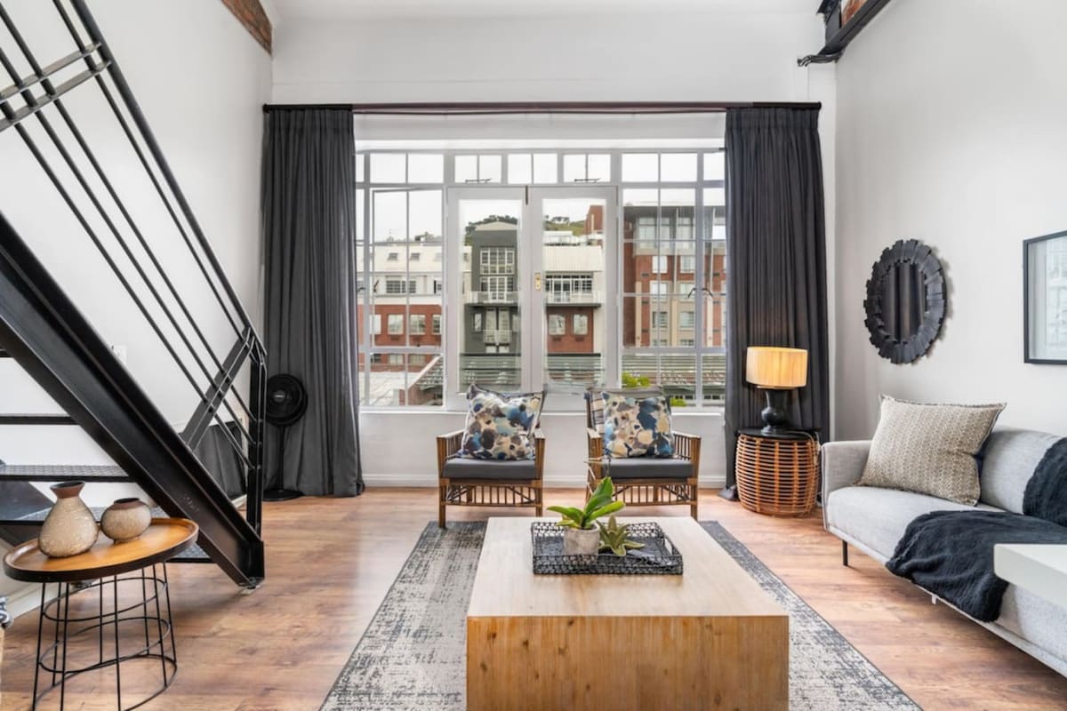 A spacious living area showcases large windows allowing ample natural light. Two comfortable armchairs with decorative pillows are complemented by a contemporary sofa. A wooden coffee table is centrally placed, while a stylish lamp and various decorative items enhance the modern aesthetic.