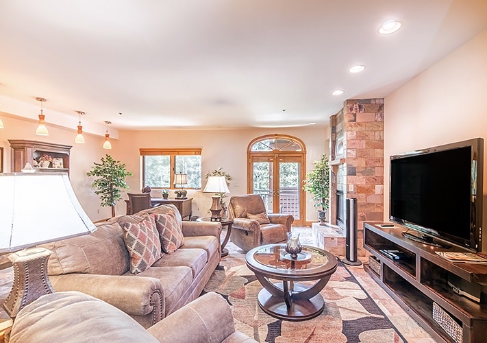 The spacious living room features soft, comfortable seating arranged around a central coffee table. A large flat screen TV is positioned against the wall, and natural light comes through the windows, enhancing the warm atmosphere. Decorative plants add a touch of greenery.