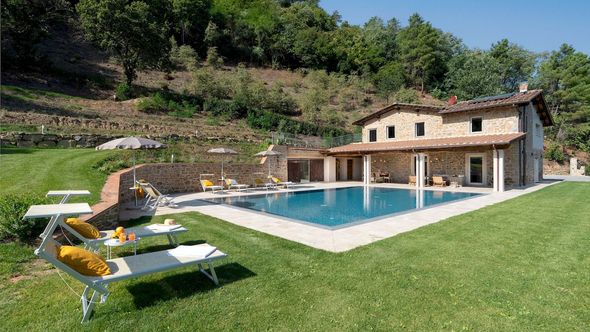 The villa's exterior showcases a rustic stone façade, surrounded by lush greenery. A rectangular swimming pool is framed by a sunbathing area equipped with lounge chairs and umbrellas. The well-maintained lawn complements the serene setting, enhancing the inviting atmosphere of the property.