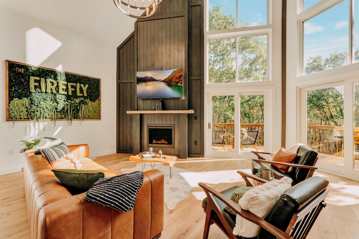 An inviting living area features a modern leather couch and comfortable seating arranged around a sleek coffee table. A wall-mounted TV sits above a contemporary fireplace, surrounded by large windows that offer abundant natural light and views of the outdoors.