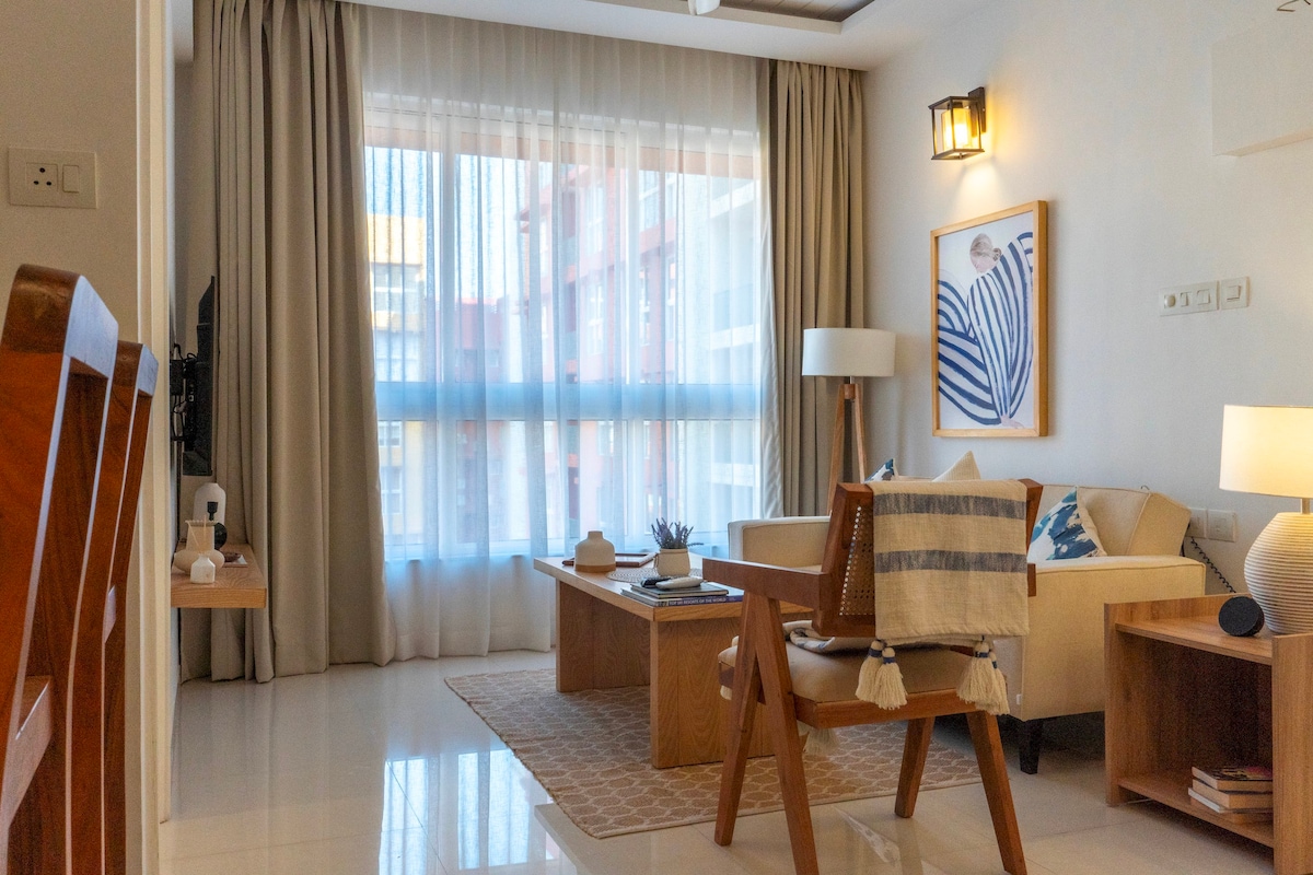 The living room offers a spacious layout with large windows allowing ample natural light. A comfortable sofa and coffee table are positioned on a light rug. An accent chair adds additional seating, while artwork adorns the wall, contributing to a calming atmosphere.
